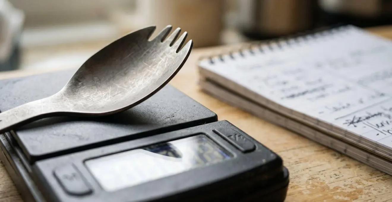 Close-up view of camping gear being weighed on a digital kitchen scale with notebook nearby for recording weights