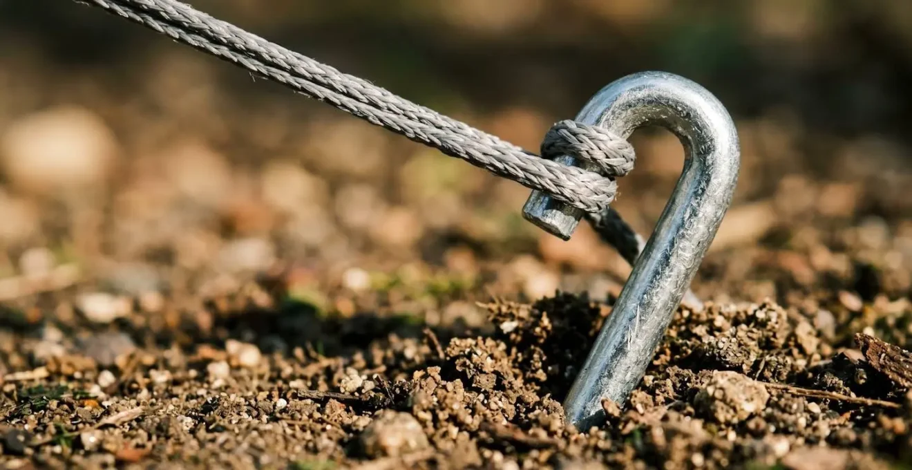 Close-up of tent peg driven into soil at precise 45-degree angle with tension cord attached